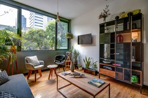 A modern living room with a large window, a sofa, a coffee table, a bookshelf, and various decorative items. - Zen Modern by Mr.W (Mexico City)