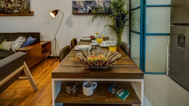 Un coin repas avec une table en bois, des chaises, une machine à café, une plante et un tableau représentant un paysage urbain accroché au mur.