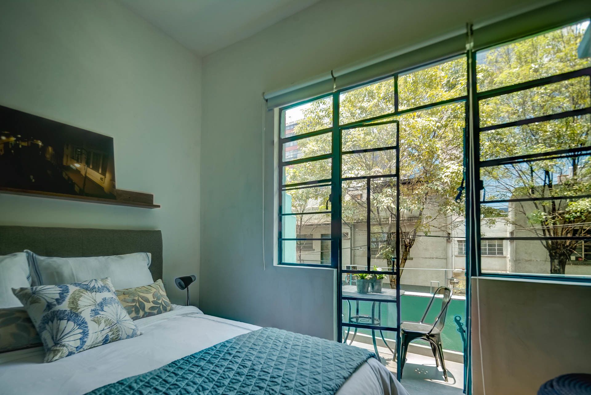 Ein Schlafzimmer mit großem Fenster, einem Bett mit gemusterten Kissen, einem Nachttisch und einem Bild an der Wand.