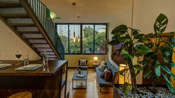 Una sala de estar moderna con una escalera de madera, un área de comedor con una mesa para dos personas y una gran planta en maceta.