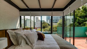 Una habitación de hotel moderna con una cama grande, vistas de la ciudad a través de ventanales de piso a techo y un balcón con valla verde.