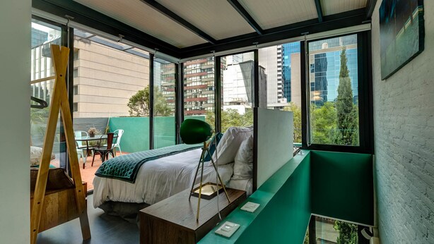 Une chambre moderne avec un lit, un bureau et une chaise. Une fenêtre offre une vue sur les bâtiments et les arbres.
