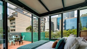 Une chambre d’hôtel moderne dotée d’un grand lit, d’un balcon avec une table et des chaises, et offrant une vue sur la ville grâce à de grandes fenêtres du sol au plafond.