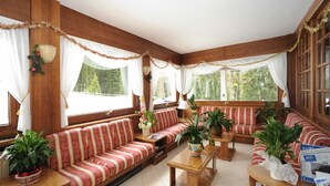 A cozy room with wooden walls and large windows, featuring a red and white striped sofa, a wooden table, and several potted plants. - Hotel Bucaneve (Moena)