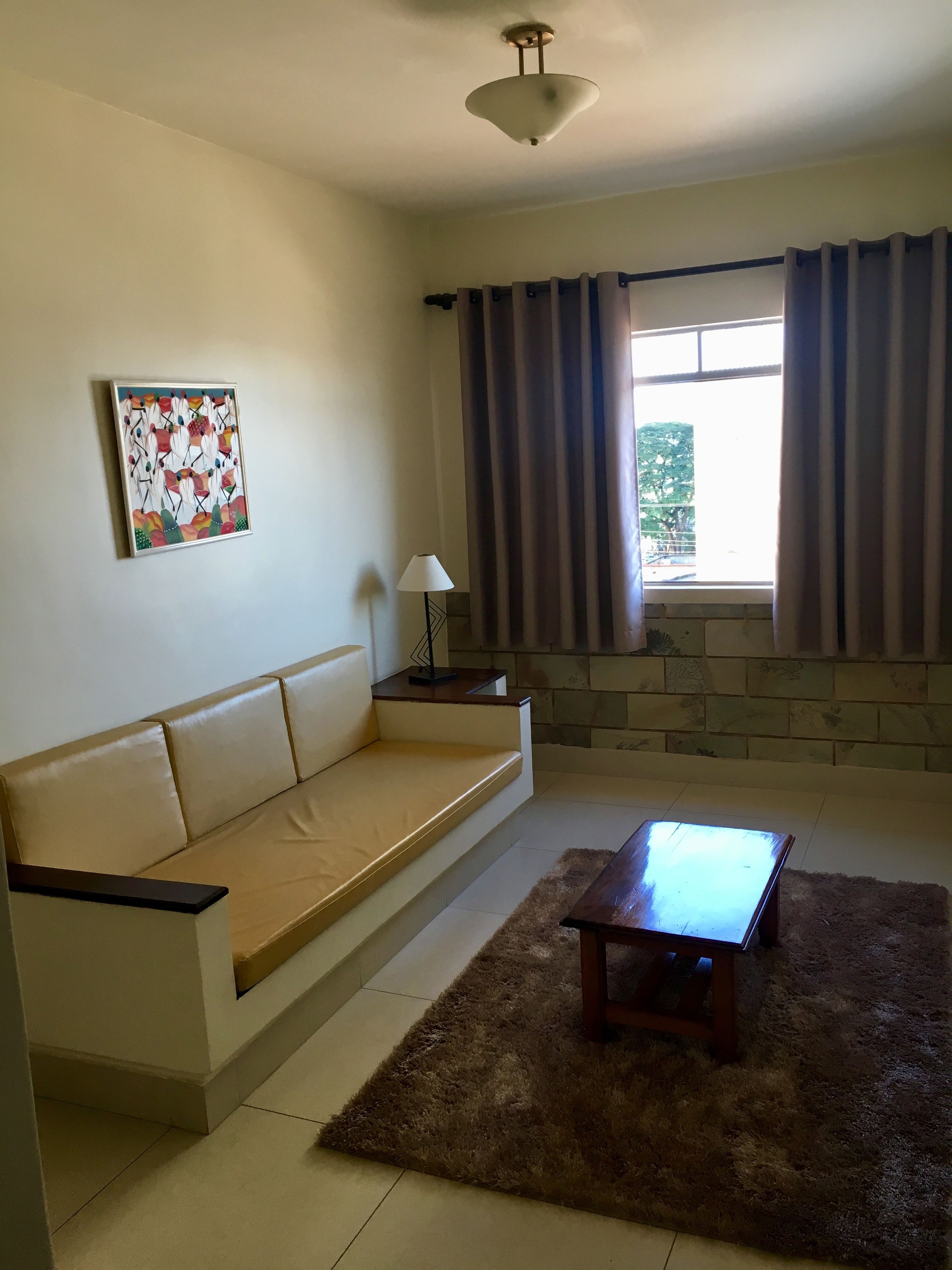 Una sala de estar con un sofá color crema, una mesa de centro de madera, una lámpara, un cuadro en la pared y un ventanal con cortinas.