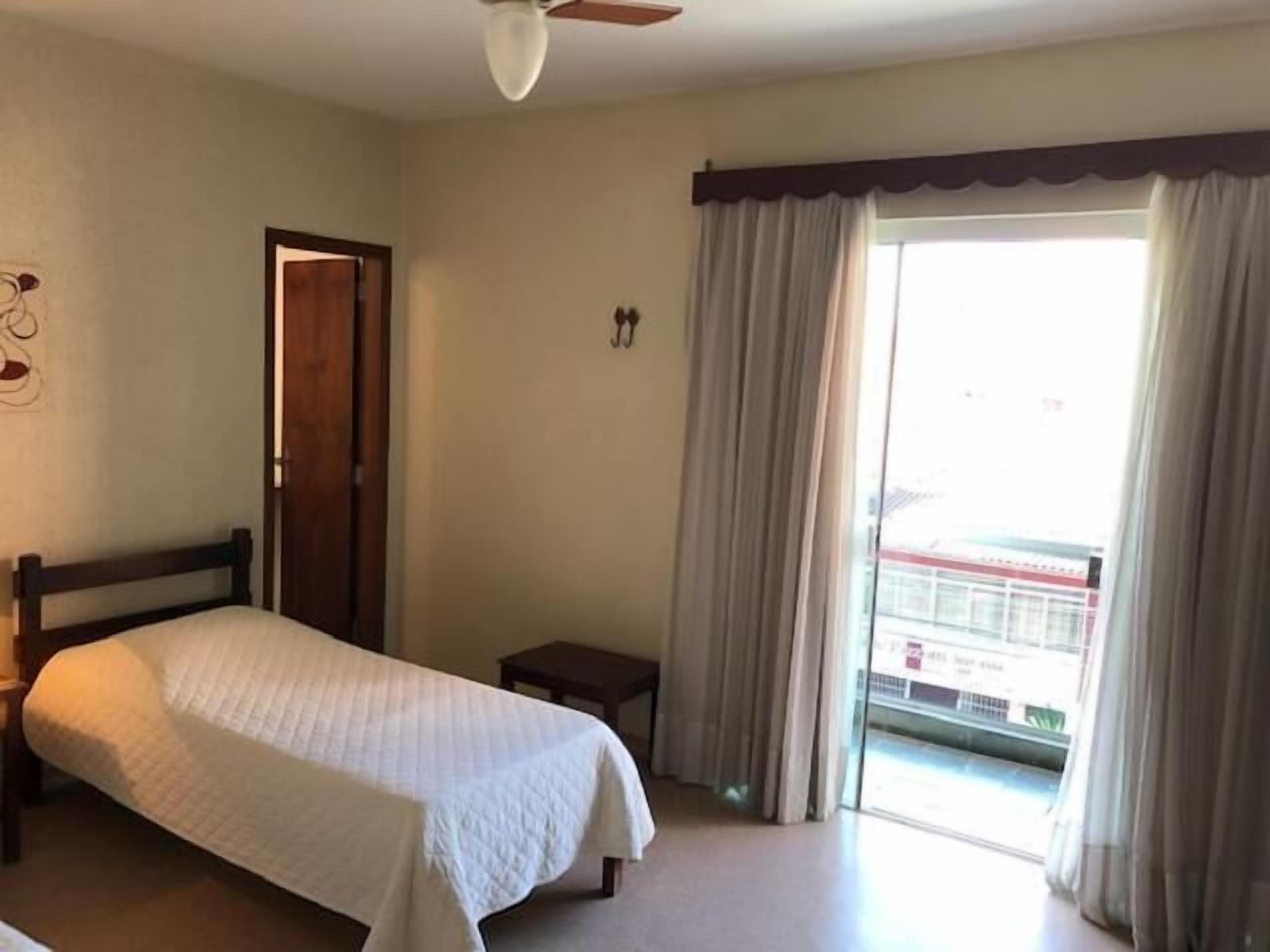 Ett hotellrum med en säng, ett litet sängbord, en takfläkt och utsikt över omgivningarna genom ett stort fönster med gardiner.
