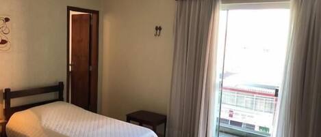 A hotel room with a bed, a small bedside table, a ceiling fan, and a view of the outside through a large window with curtains.