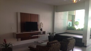 Een moderne woonkamer met een flatscreen televisie, twee fauteuils, een salontafel en een groot raam met uitzicht op groen.