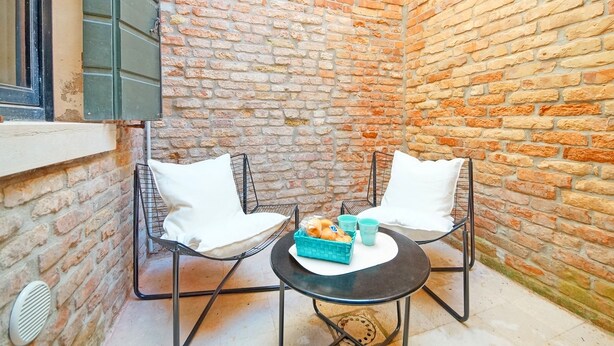 A small, enclosed patio area with brick walls, two white cushioned chairs, a round table with a basket of bread and two cups, and a window with blinds.