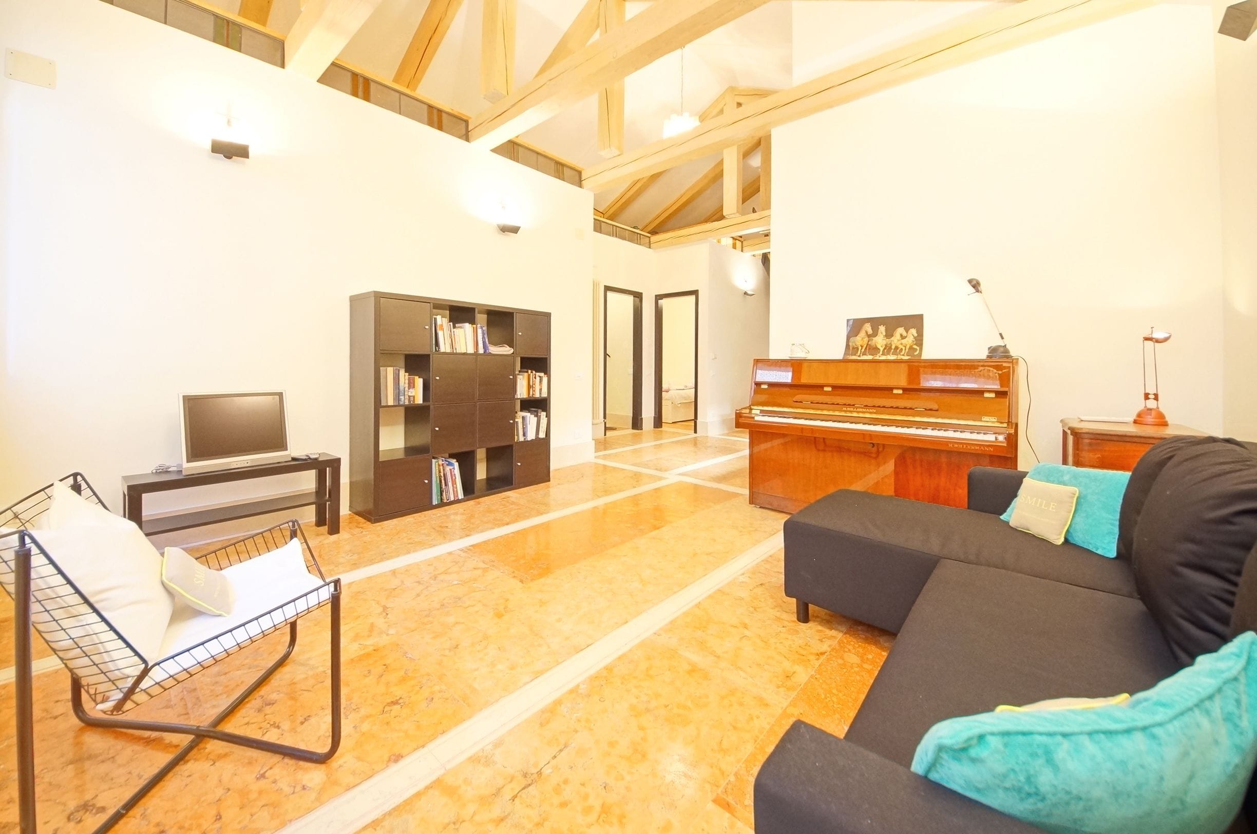 A spacious living room with a black sofa, a metal chair, a bookshelf, a piano, and a television.