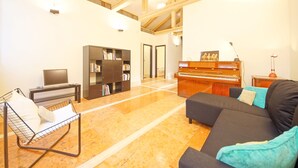 A spacious living room with a black sofa, a metal chair, a bookshelf, a piano, and a television.