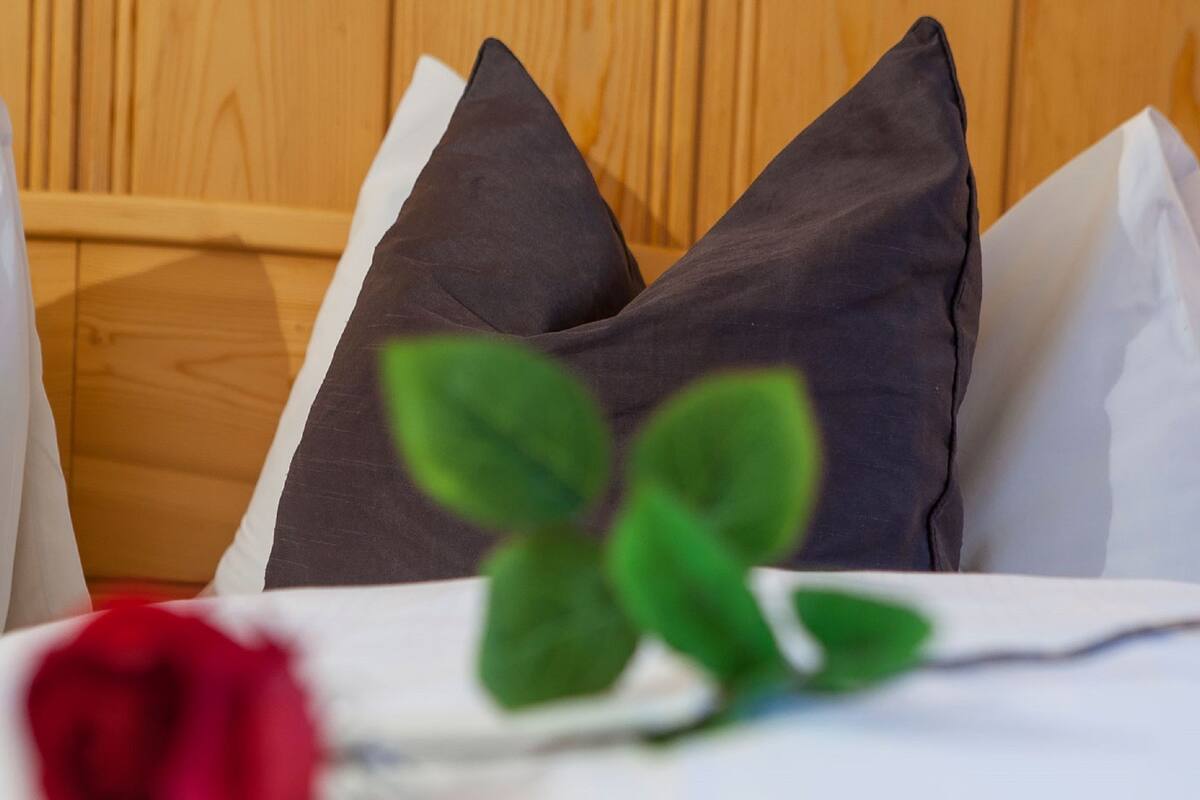 A bed with white and dark pillows, a red rose, and green leaves.