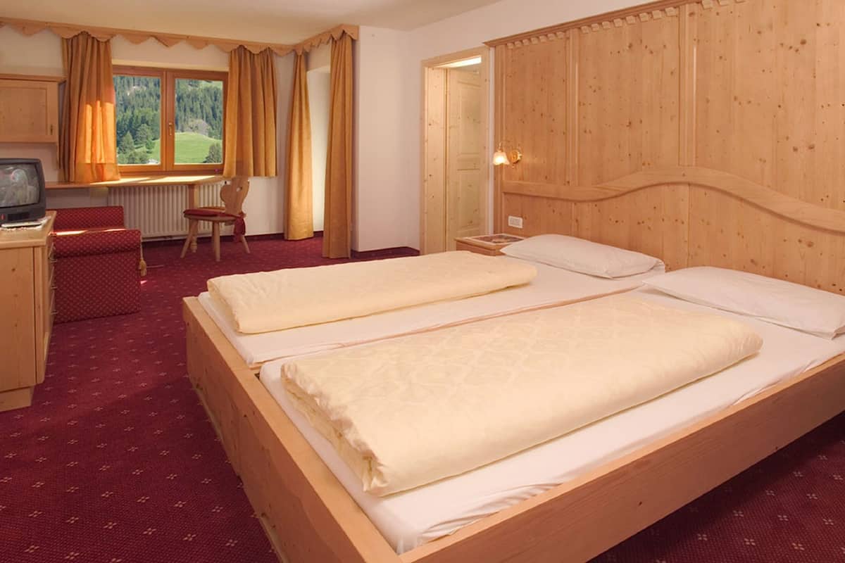 A hotel room with a wooden headboard, two beds with white linens, a wooden desk with a television, a chair, and a window with curtains.