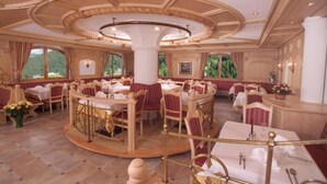 A spacious dining area with round tables and chairs, large windows, and a central column. - Hotel Jagdhof (Selva di Val Gardena)