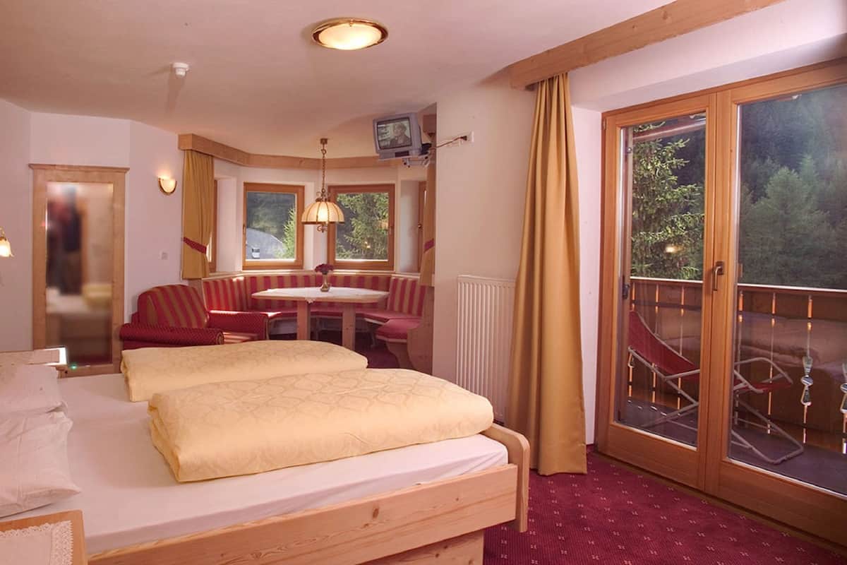 A hotel room with a wooden headboard, two beds with beige linens, a small table with chairs, a balcony with a glass railing, and a television mounted on the wall.