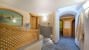 A hallway with a wooden door, a wall-mounted light fixture, and a stone floor. - Hotel Jagdhof (Selva di Val Gardena)