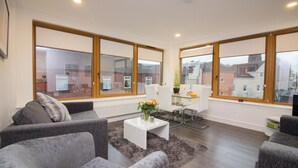 Una sala de estar moderna con sofás grises, una mesa de centro blanca y una mesa de comedor de vidrio. Amplias ventanas ofrecen vistas de edificios.