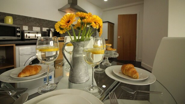 Una mesa de comedor con platos, cruasanes, copas de vino y un florero con flores amarillas.
