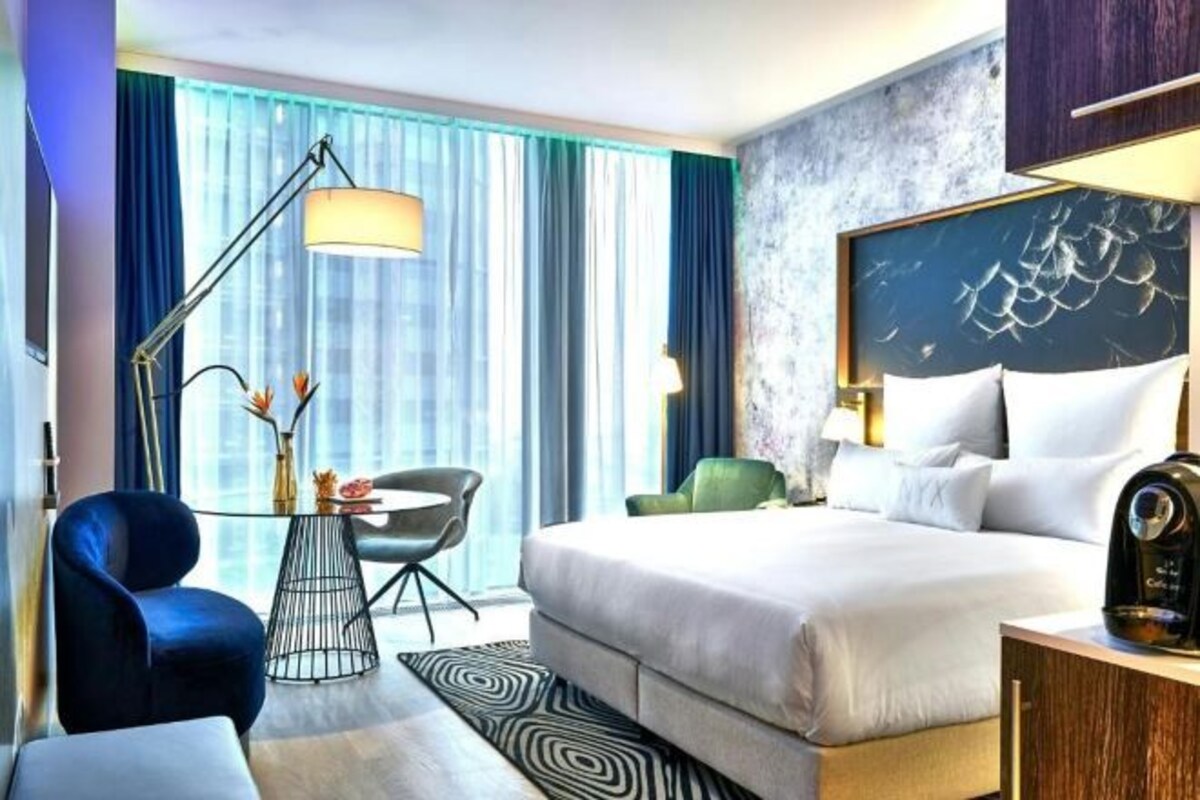 Habitación de hotel con cama, escritorio con silla, lámpara y vistas a la ciudad a través de amplios ventanales.