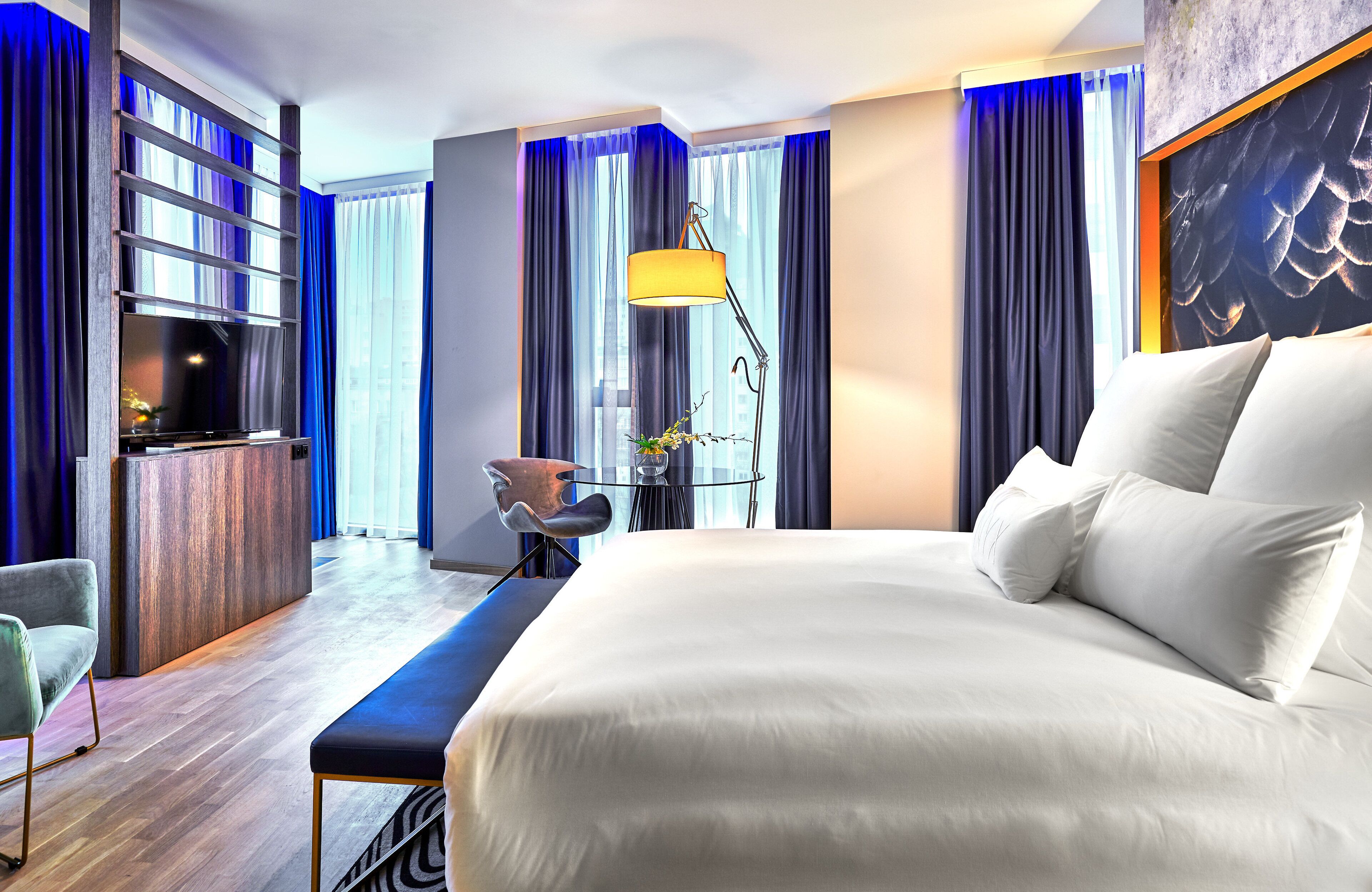 A modern hotel room with a large bed, a desk, a chair, and a floor lamp.