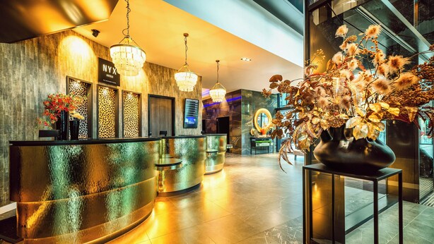 A hotel reception area with a curved reception desk, chandeliers, a digital display, and a large floral arrangement. - NYX Hotel Warsaw by Leonardo Hotels (Warsaw)