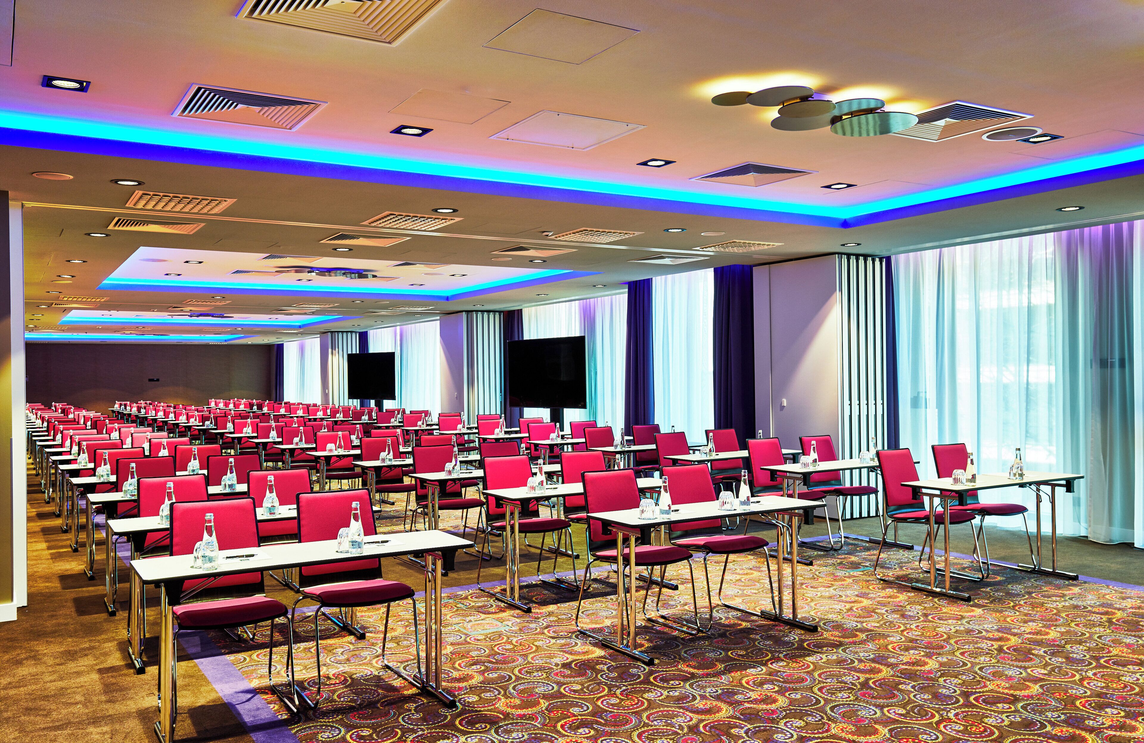 A conference room with rows of tables and chairs, equipped with water bottles and nameplates, and large windows with curtains.