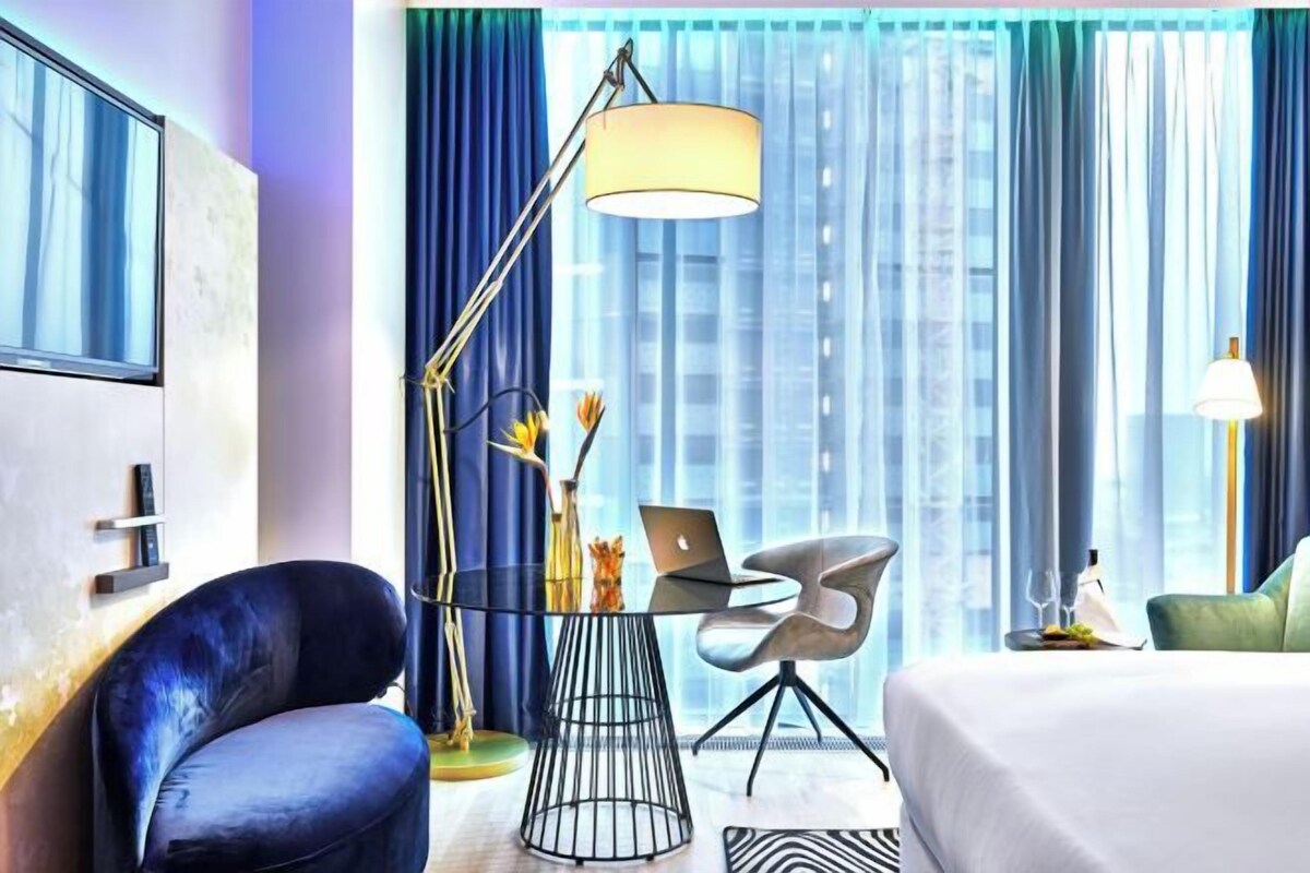 A modern hotel room with a large bed, a desk with a laptop, a chair, and a floor lamp.