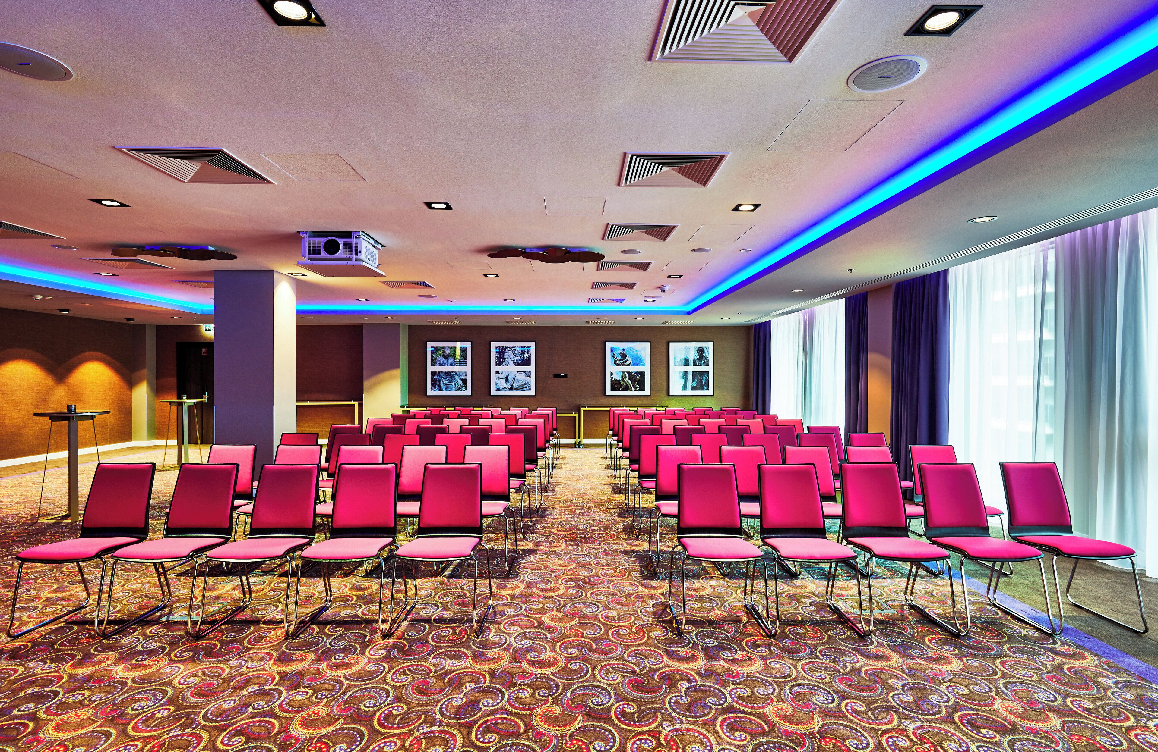 A conference room with rows of red chairs, a patterned carpet, and ceiling lights.