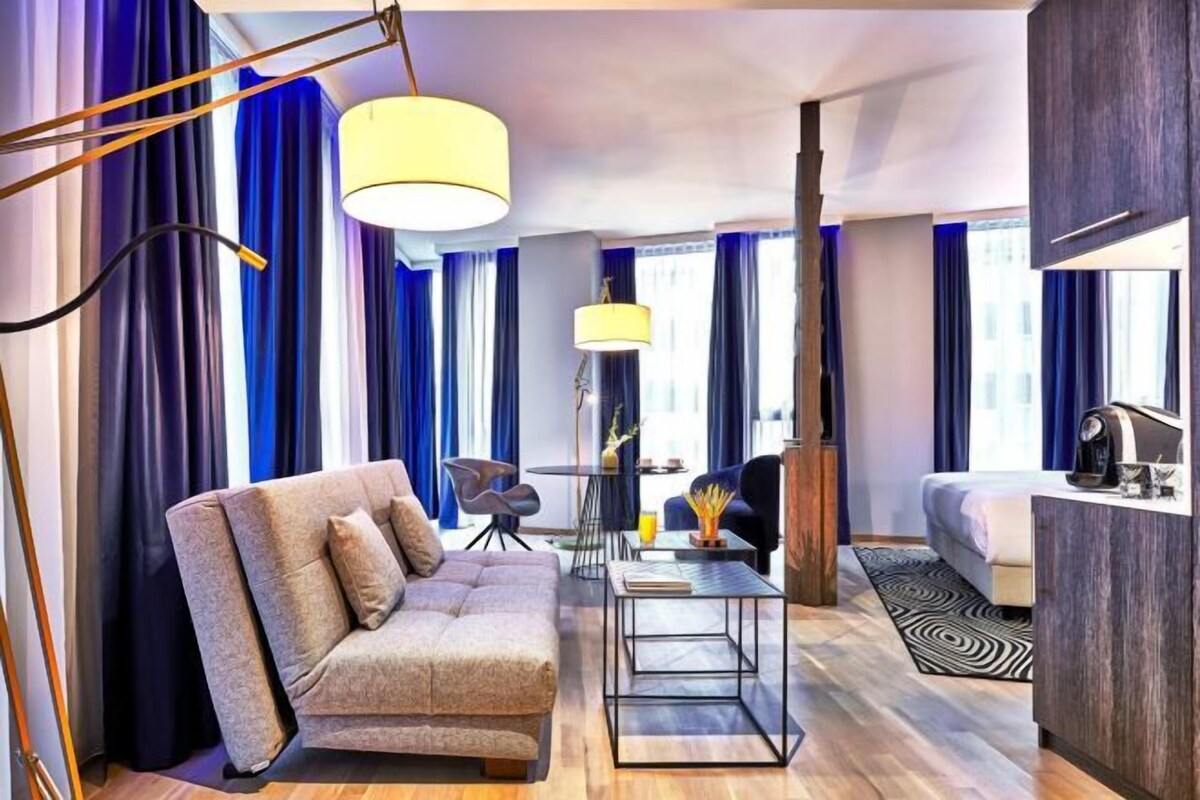 Una habitación de hotel moderna con sofá, mesa de comedor, cama y un amplio ventanal con cortinas.