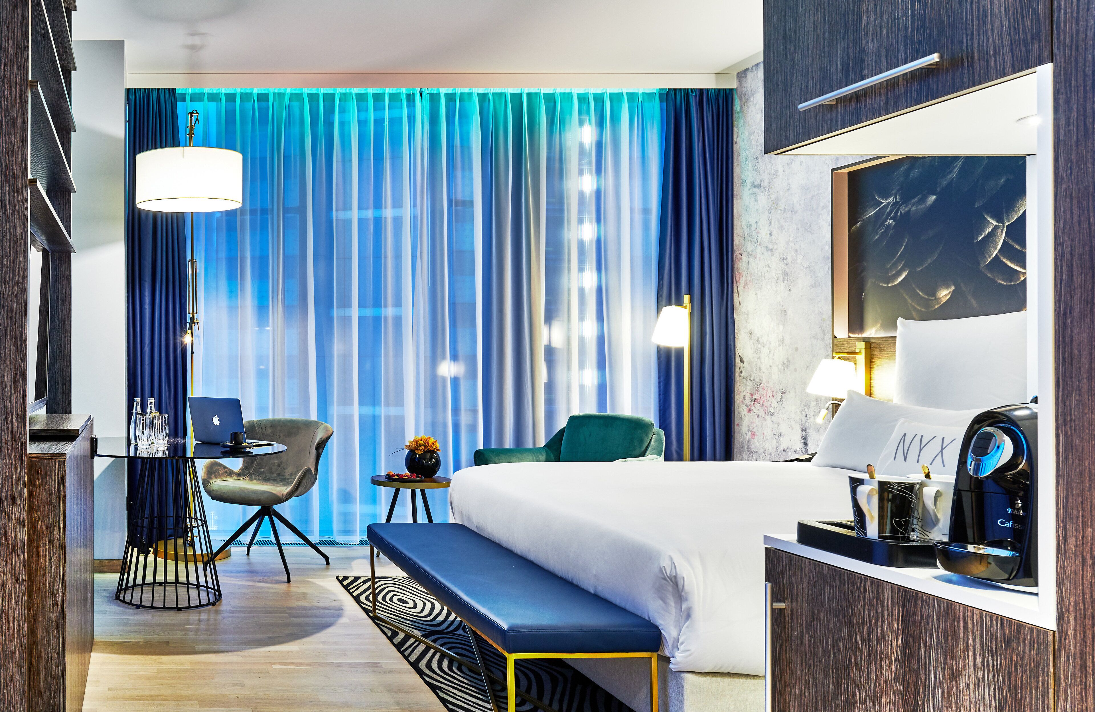 A modern hotel room with a large bed, a desk with a laptop, a chair, a bench, and a view of the city through sheer curtains.