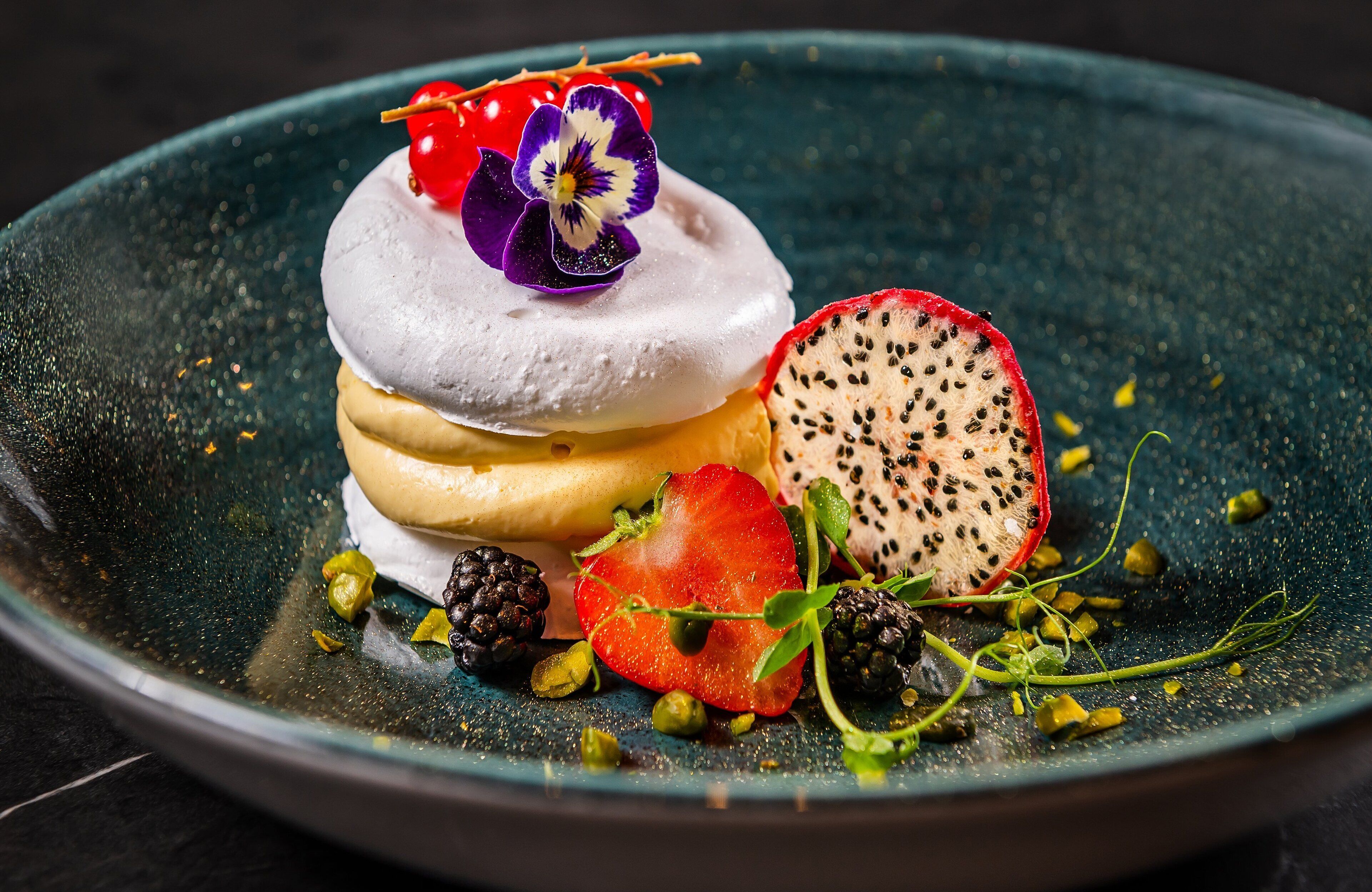 Un postre con merengue, fresas, zarzamoras y una flor decorativa sobre un plato oscuro.