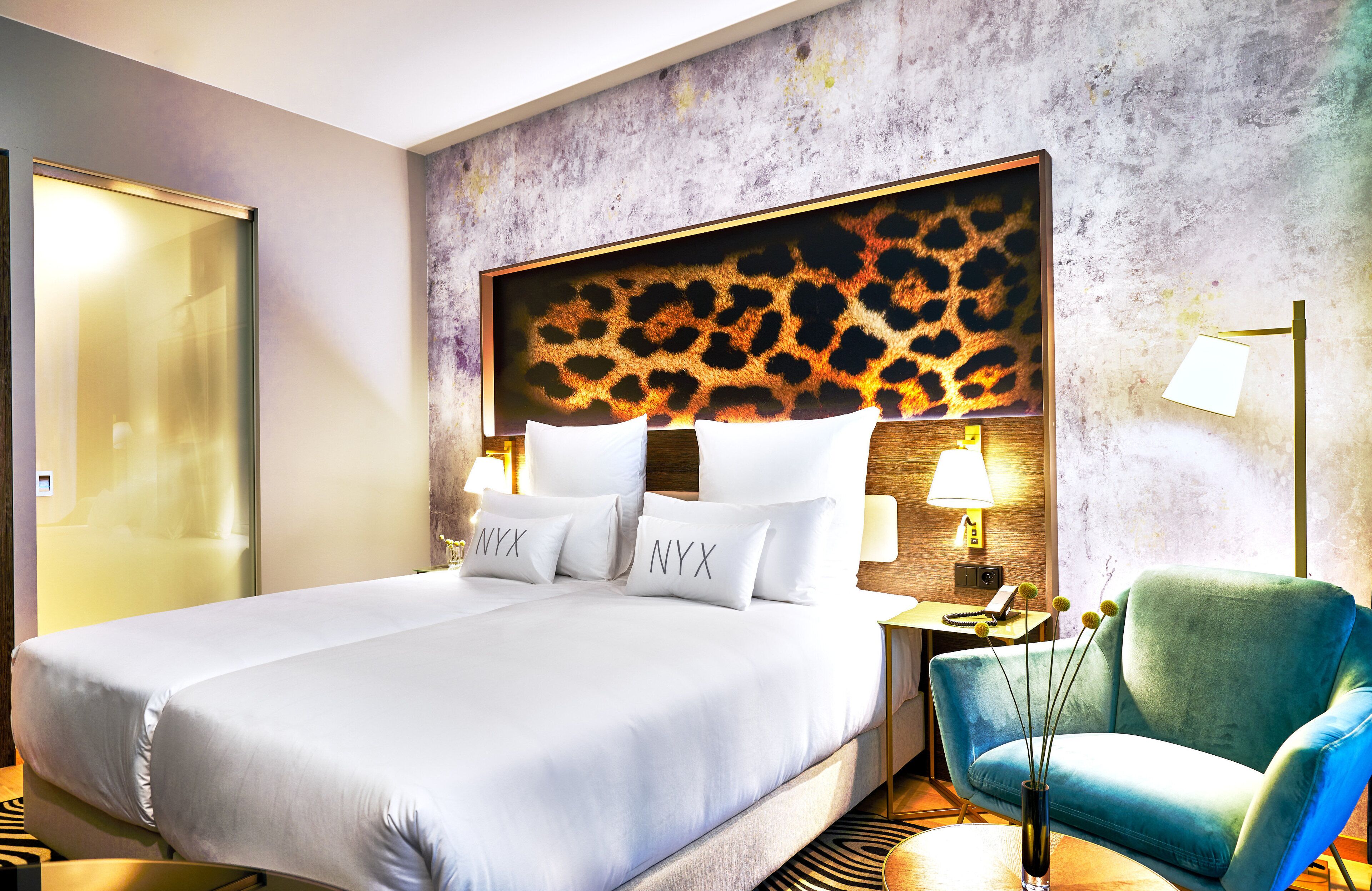 Una habitación de hotel con una cama grande, un cabecero estampado de leopardo, dos lámparas de noche, un sillón verde y una mesita con un florero.