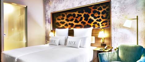 Una habitación de hotel con una cama grande, un cabecero estampado de leopardo, dos lámparas de noche, un sillón verde y una mesita con un florero.