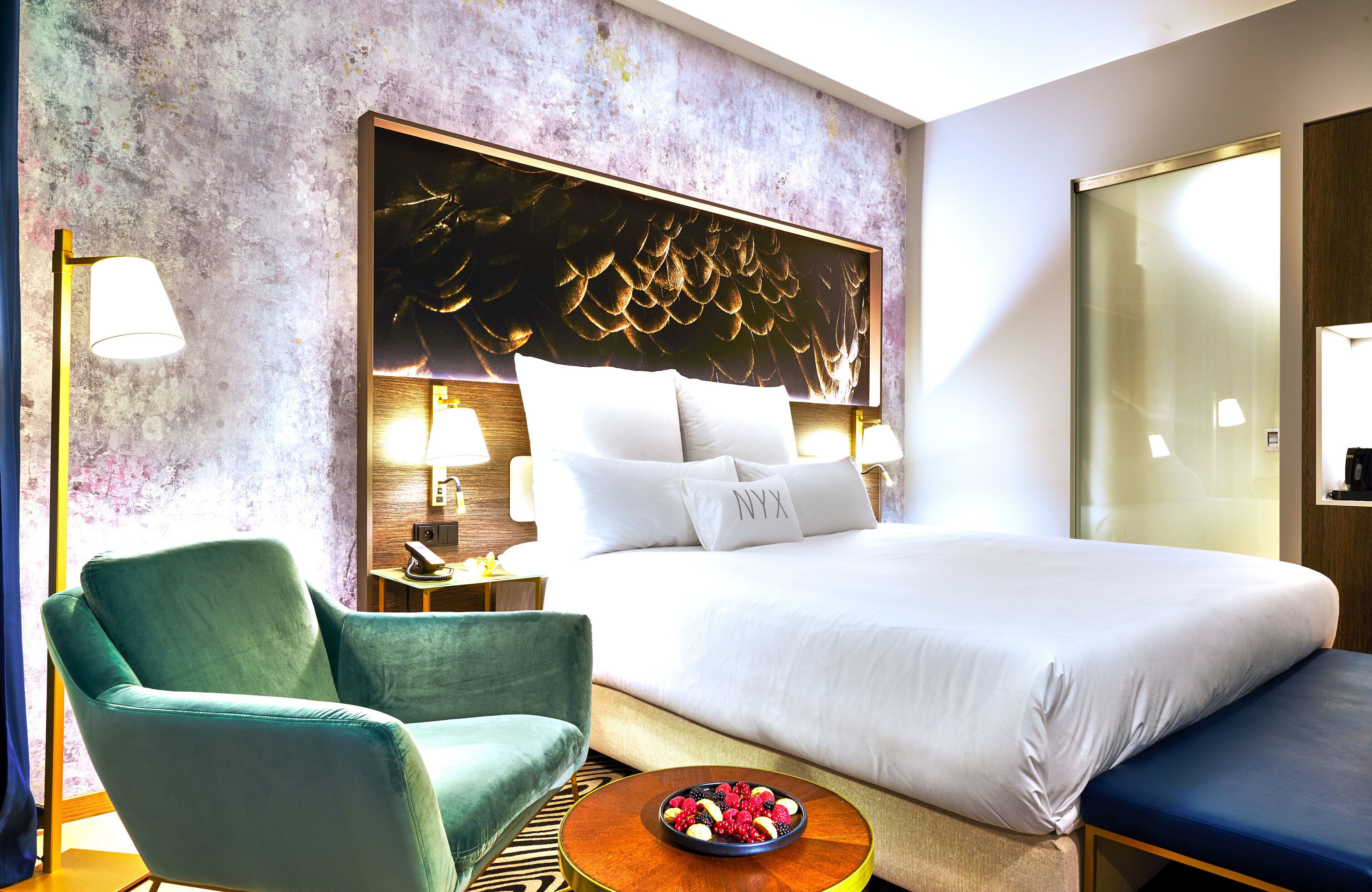 A hotel room with a large bed, a green armchair, a bedside table with a lamp, and a fruit bowl on a round table.