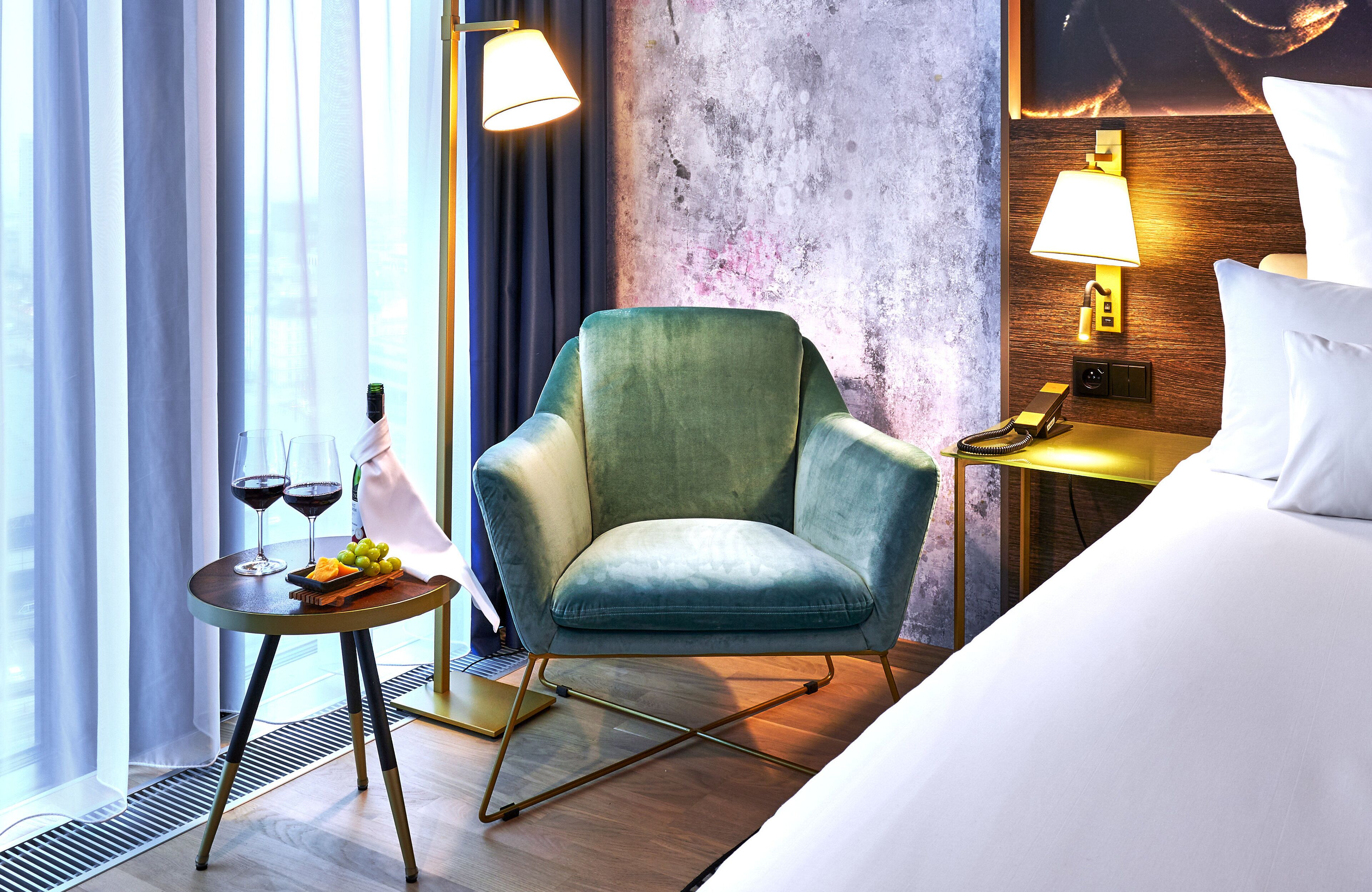 A hotel room with a green armchair, a bedside table with a lamp, a bed, a window with curtains, and a wall with a textured design.