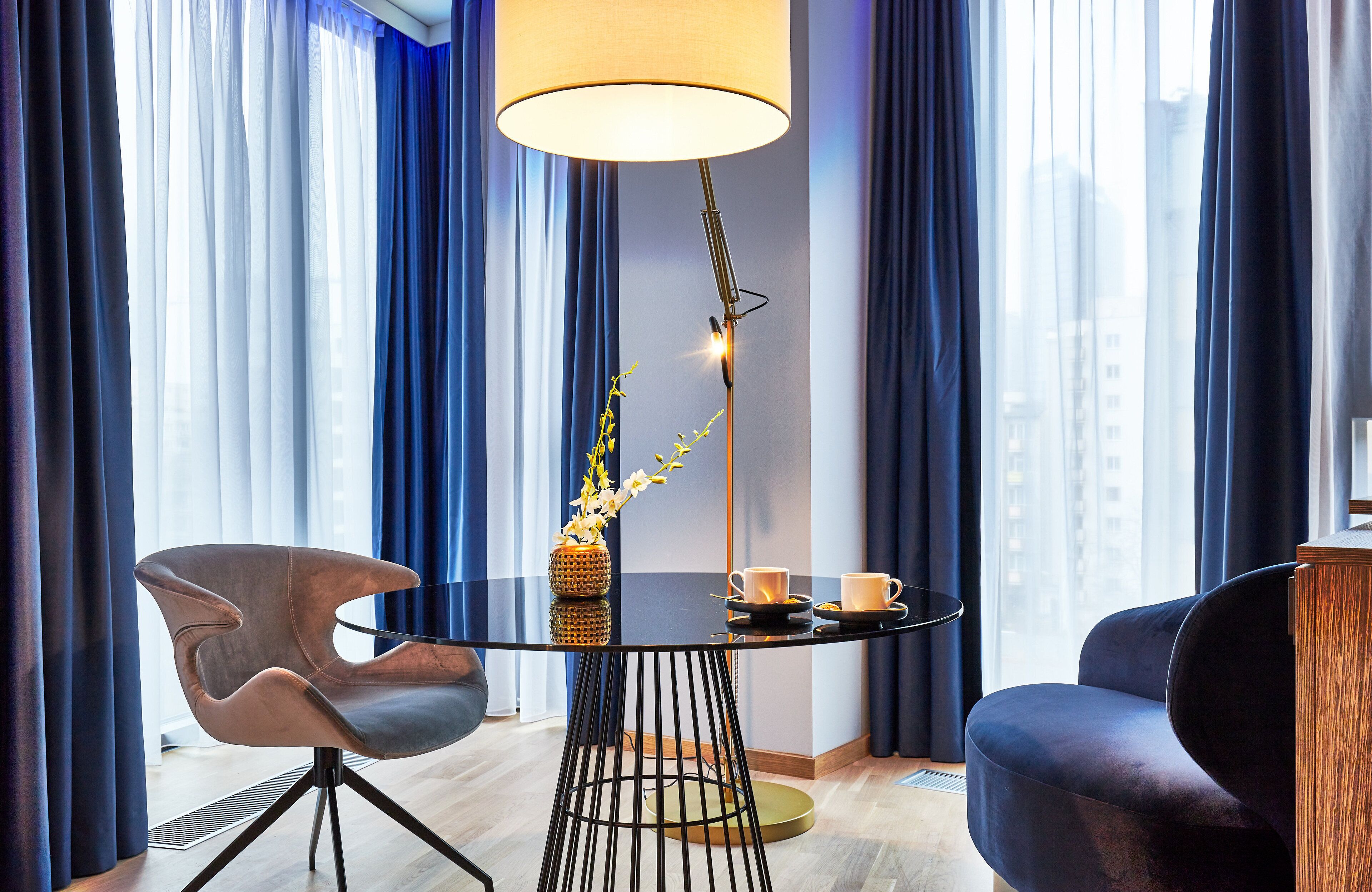 Une chambre d’hôtel moderne avec une table ronde noire, deux chaises, un lampadaire et un vase rempli de fleurs.