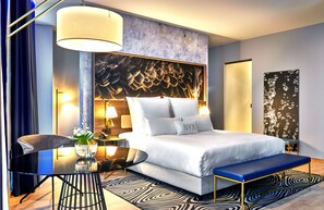 A modern hotel room with a large bed, a bedside table with a lamp, a chair, a small table with a vase of flowers, and a large abstract painting on the wall. - NYX Hotel Warsaw by Leonardo Hotels (Warsaw)