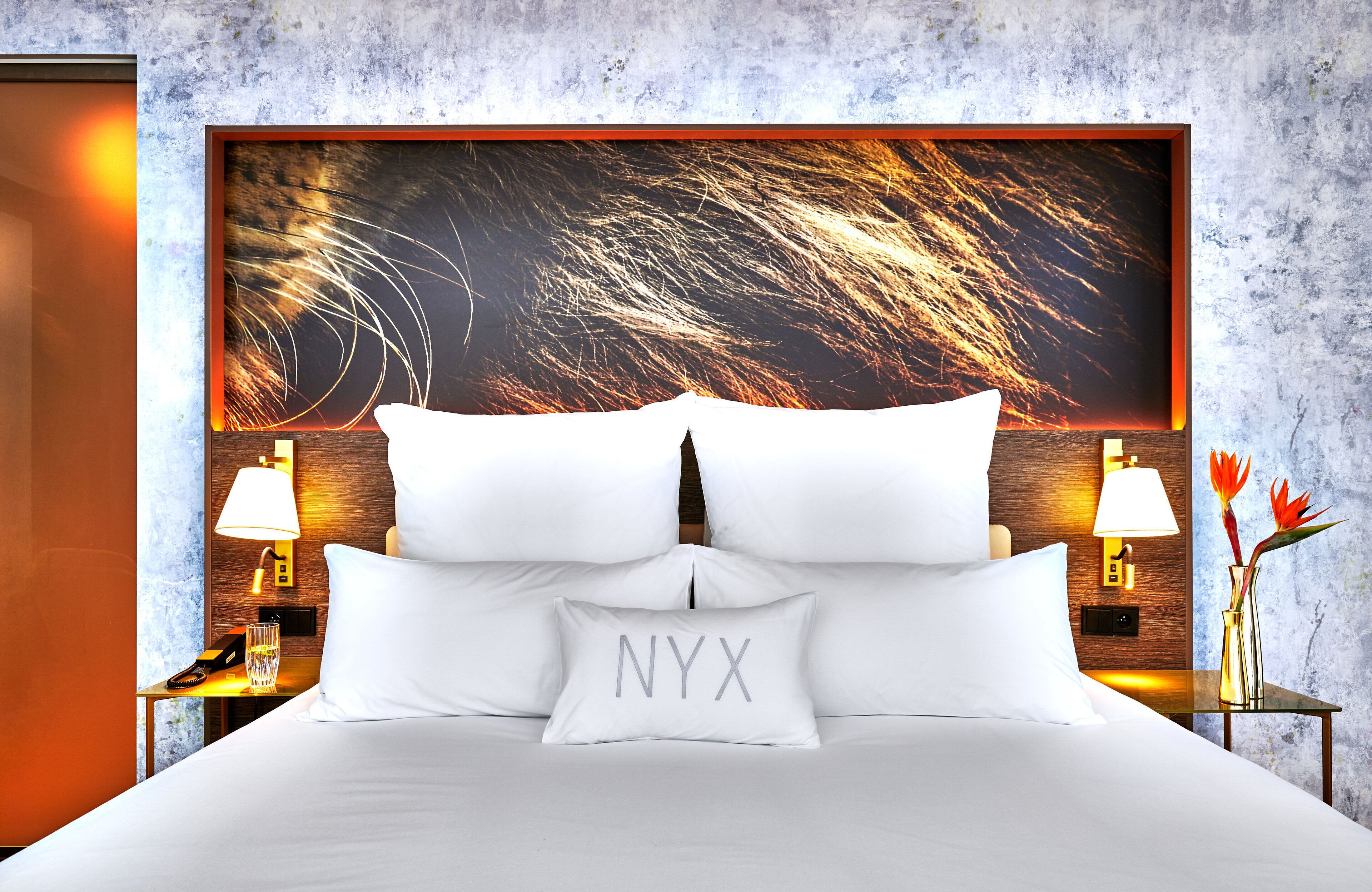 Una habitación de hotel con una cama grande, ropa de cama blanca, una obra de arte enmarcada en la cabecera, lámparas de noche, una mesita de noche con un teléfono y un jarrón con flores.
