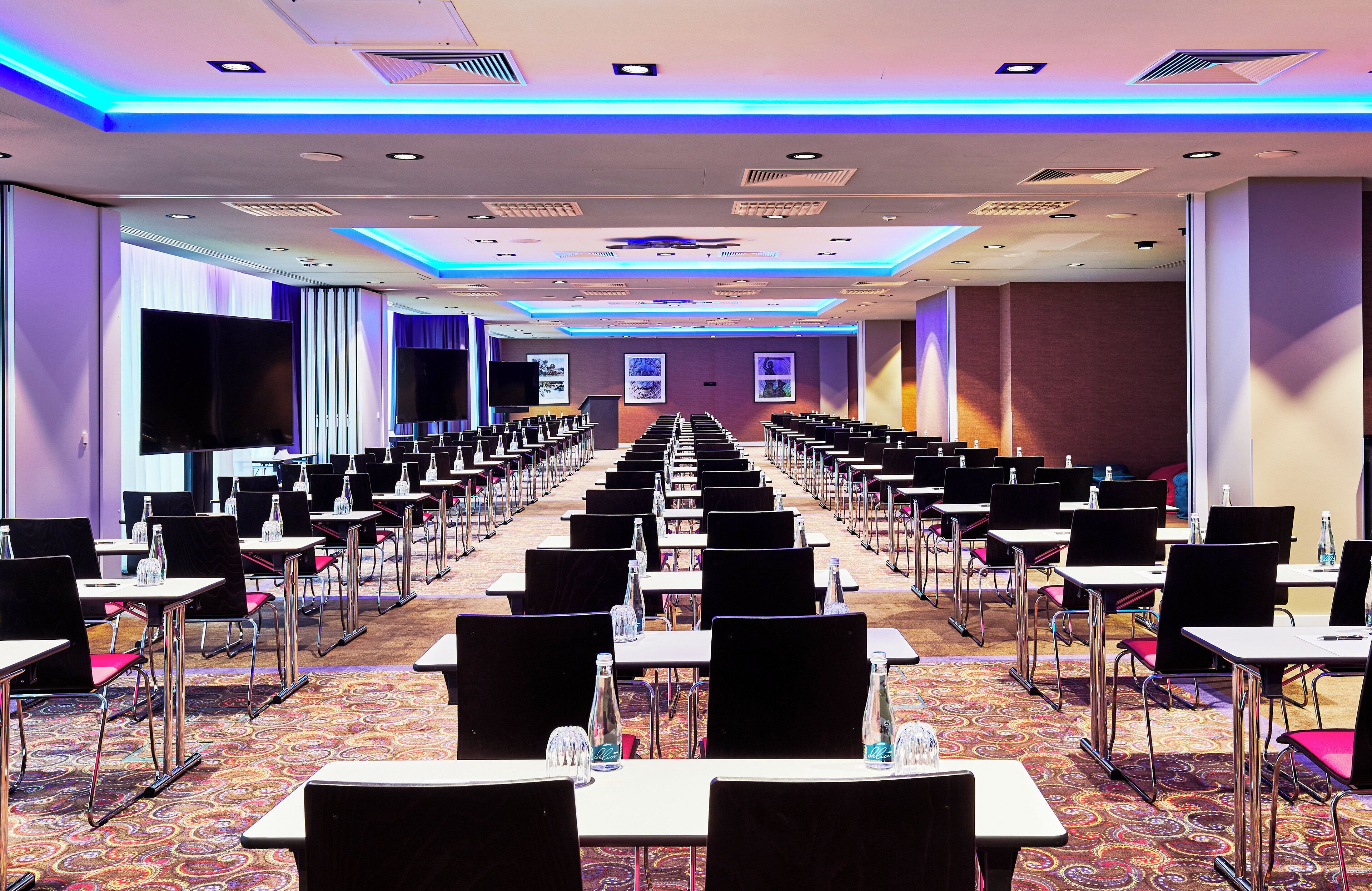 Una sala conferenze con file di tavoli e sedie, grandi schermi e pavimento con tappeto.