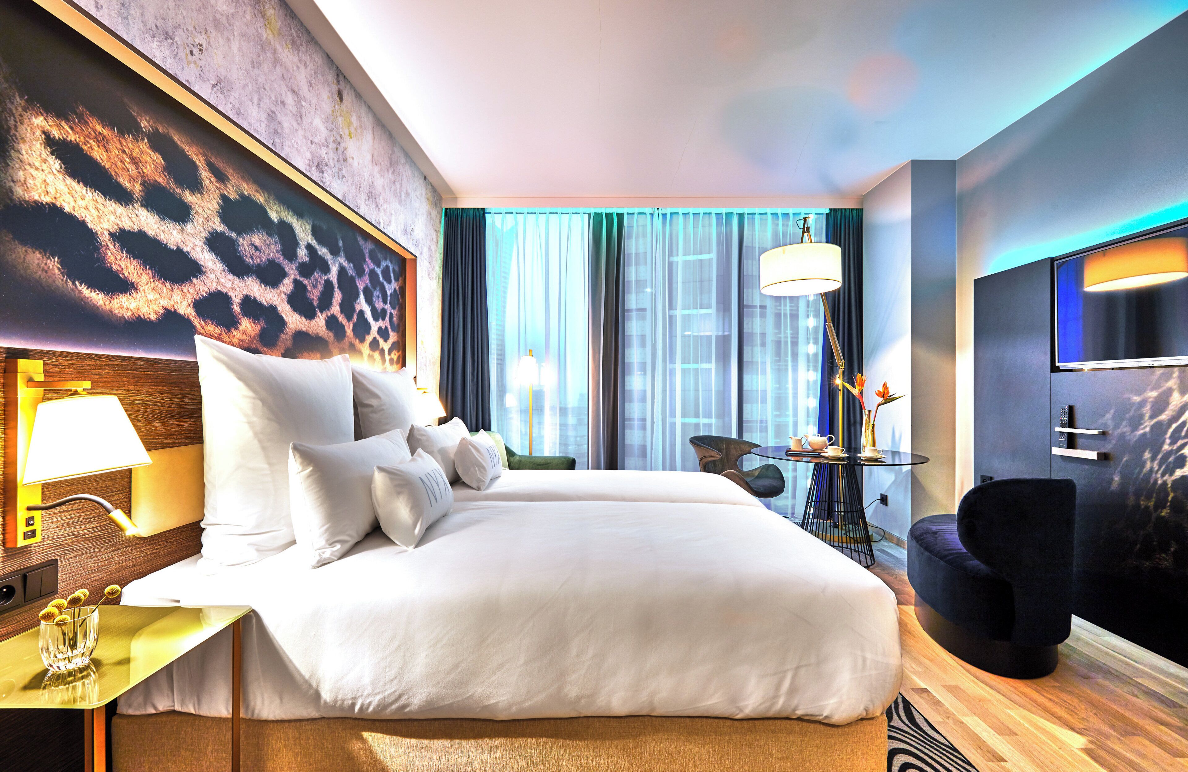 Habitación de hotel con una cama grande, un escritorio con lámpara, una silla, un televisor y vista a la ciudad por la ventana.