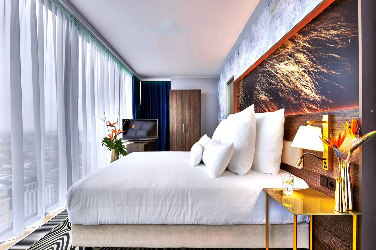 A modern hotel room with a large bed, a flat-screen TV, a wooden wardrobe, and a city view.