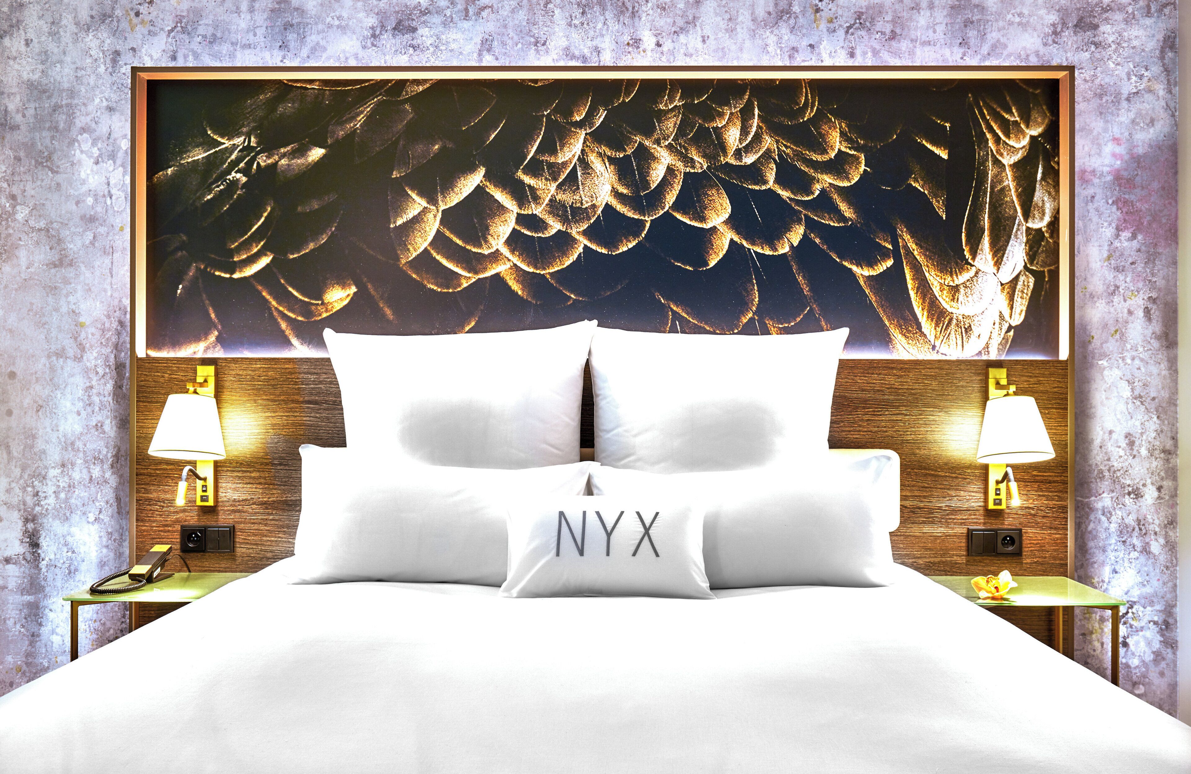 A bed with white bedding and a pillow with 'NYX' on it, flanked by two bedside tables with lamps, against a textured wall.