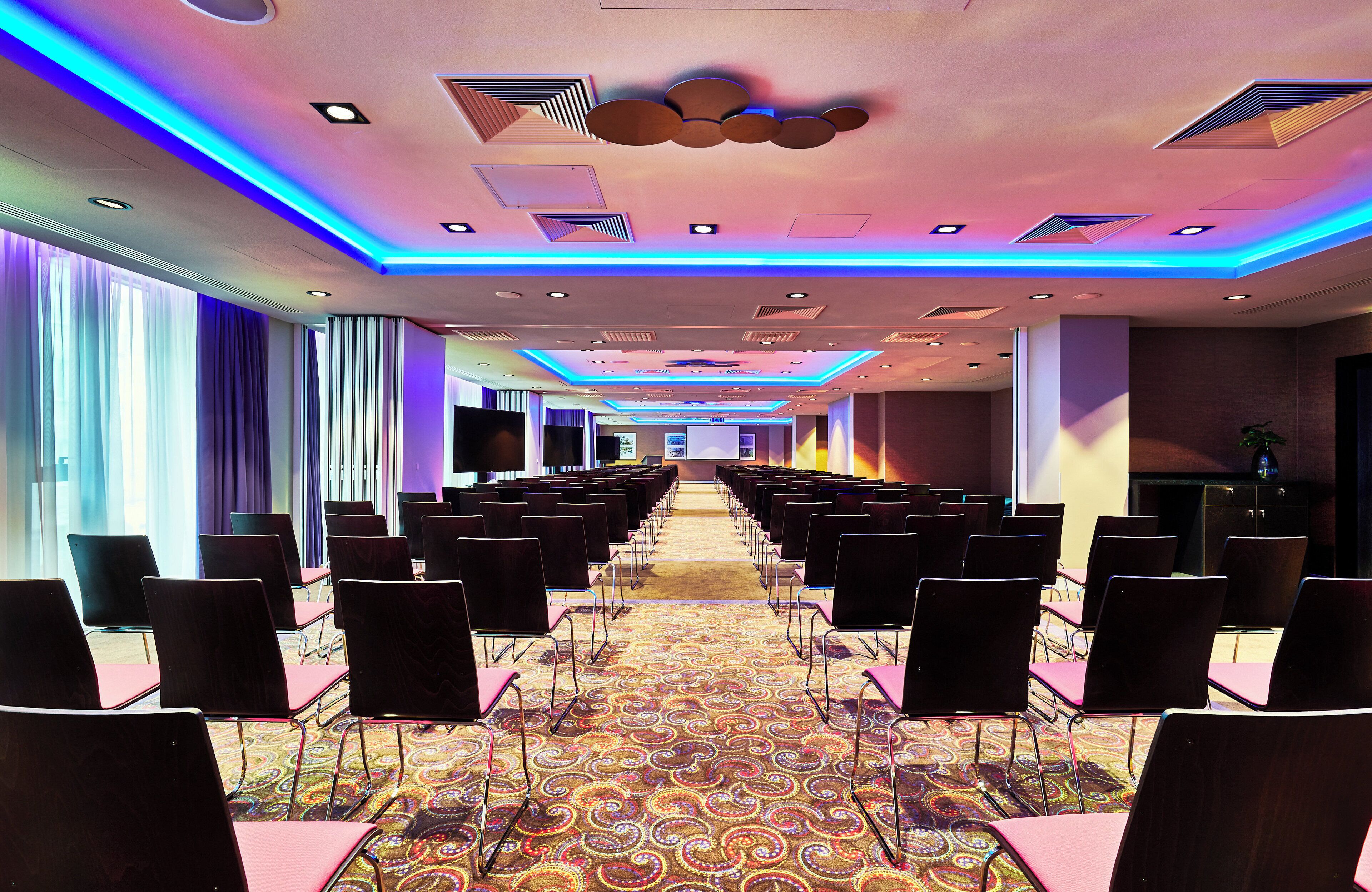 Una sala conferenze con file di sedie, una moquette fantasia e un soffitto con illuminazione a incasso e decorazioni.