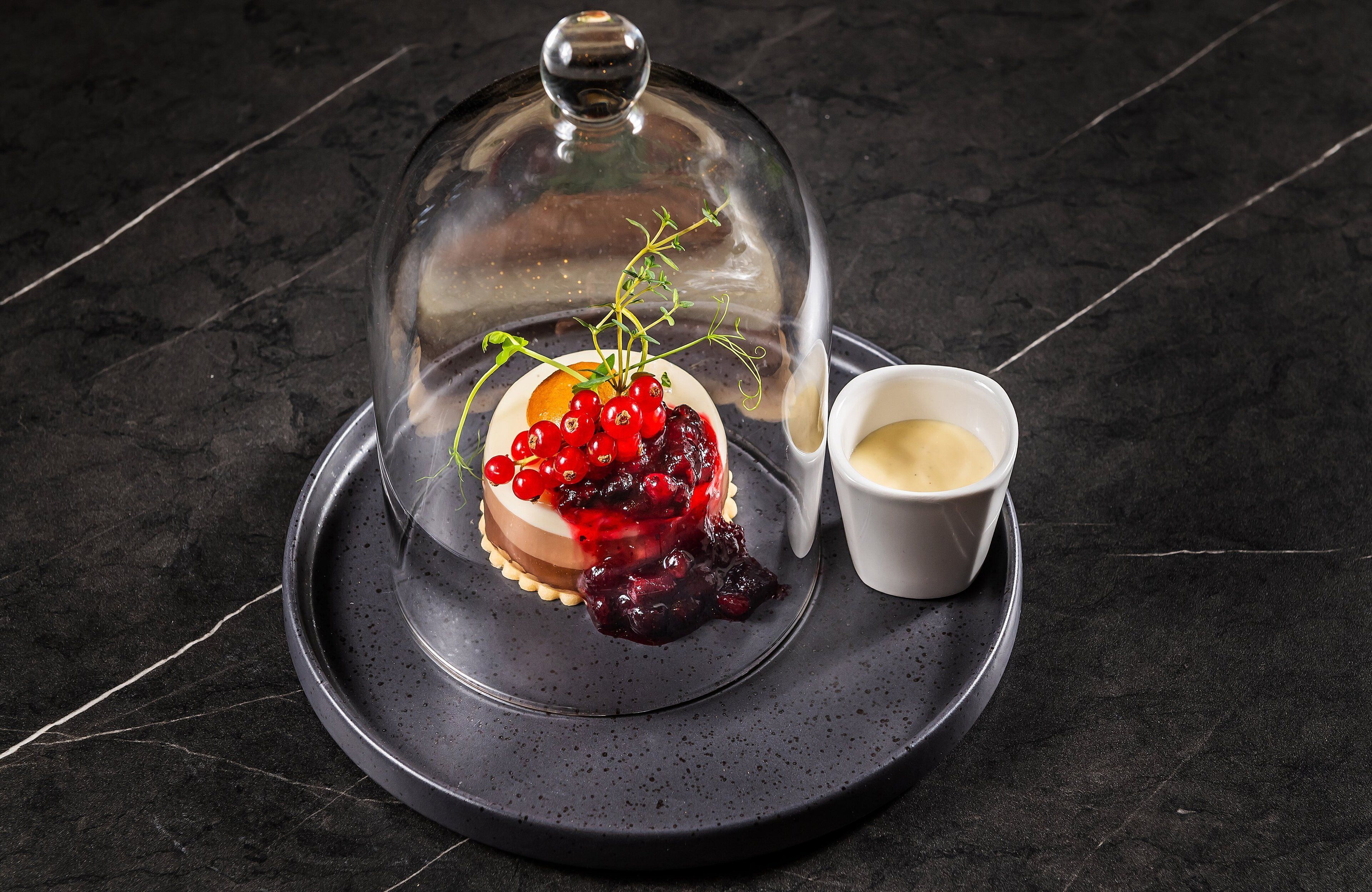 Sobremesa com cúpula de vidro sobre um prato, acompanhada de uma pequena taça de creme.