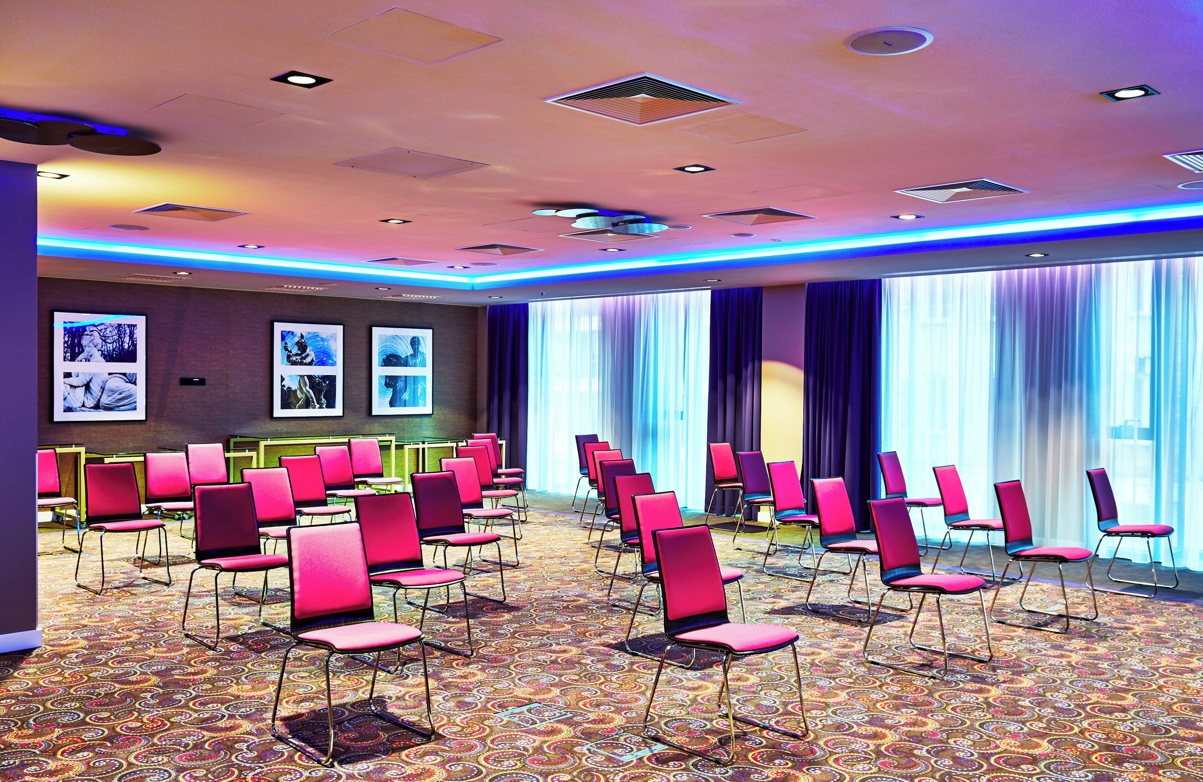 A conference room with rows of pink chairs, a patterned carpet, and large windows with curtains.