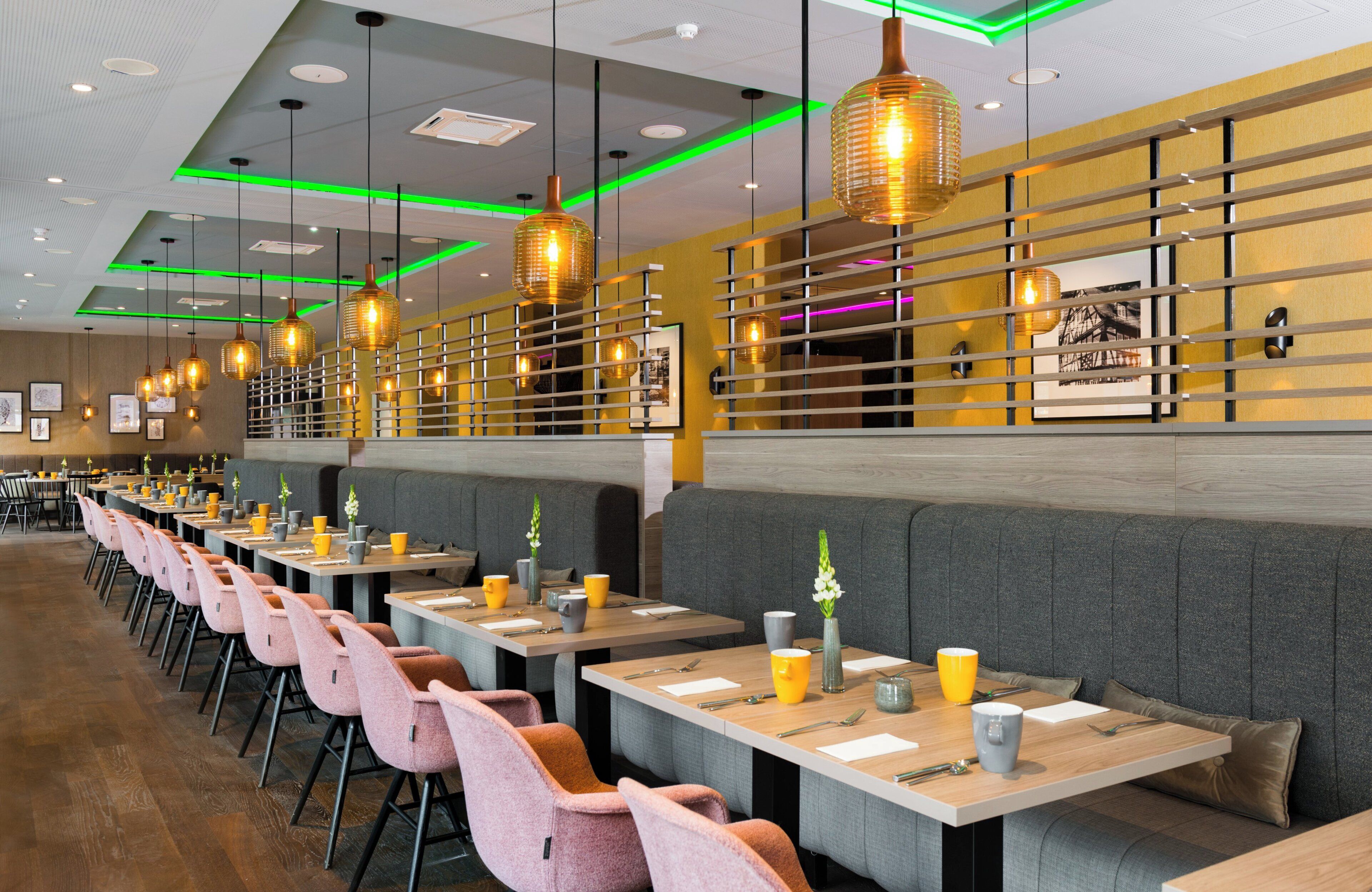 Interior de restaurante moderno com mesas compridas, cabines estofadas e iluminação pendente.