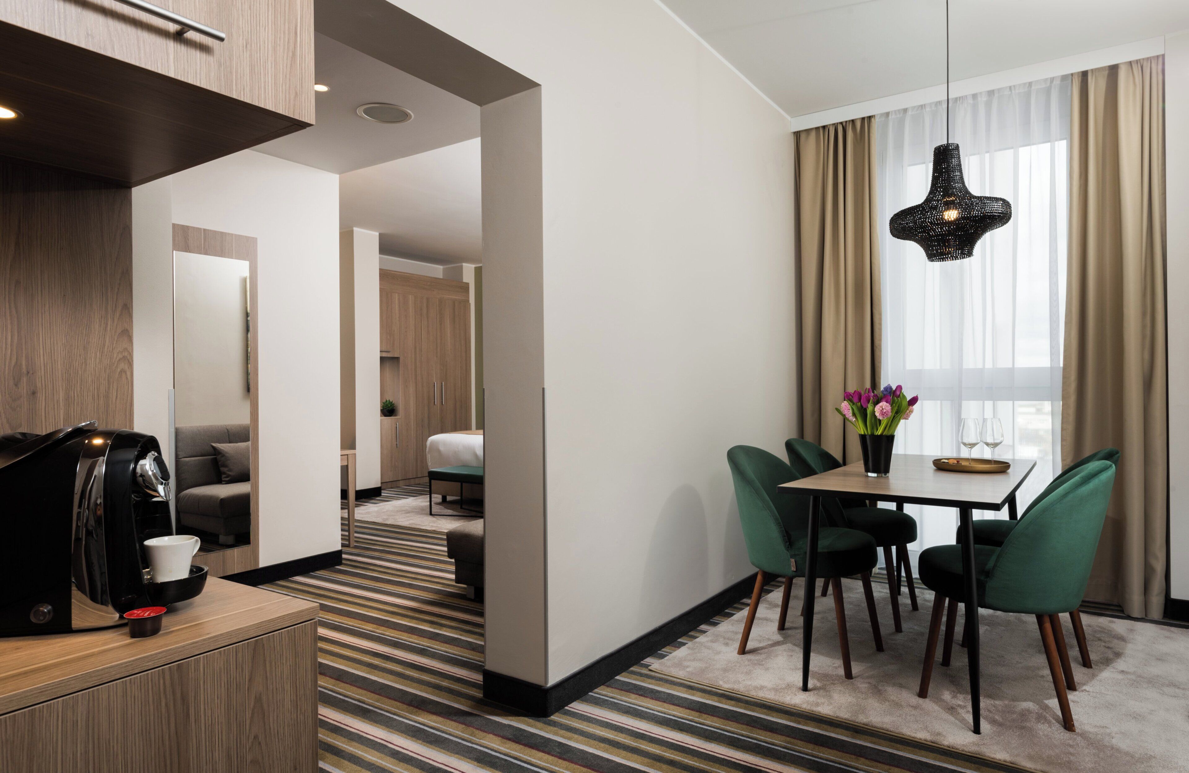 A hotel room with a dining area, a coffee machine, a sofa, a bed, and a window with curtains.