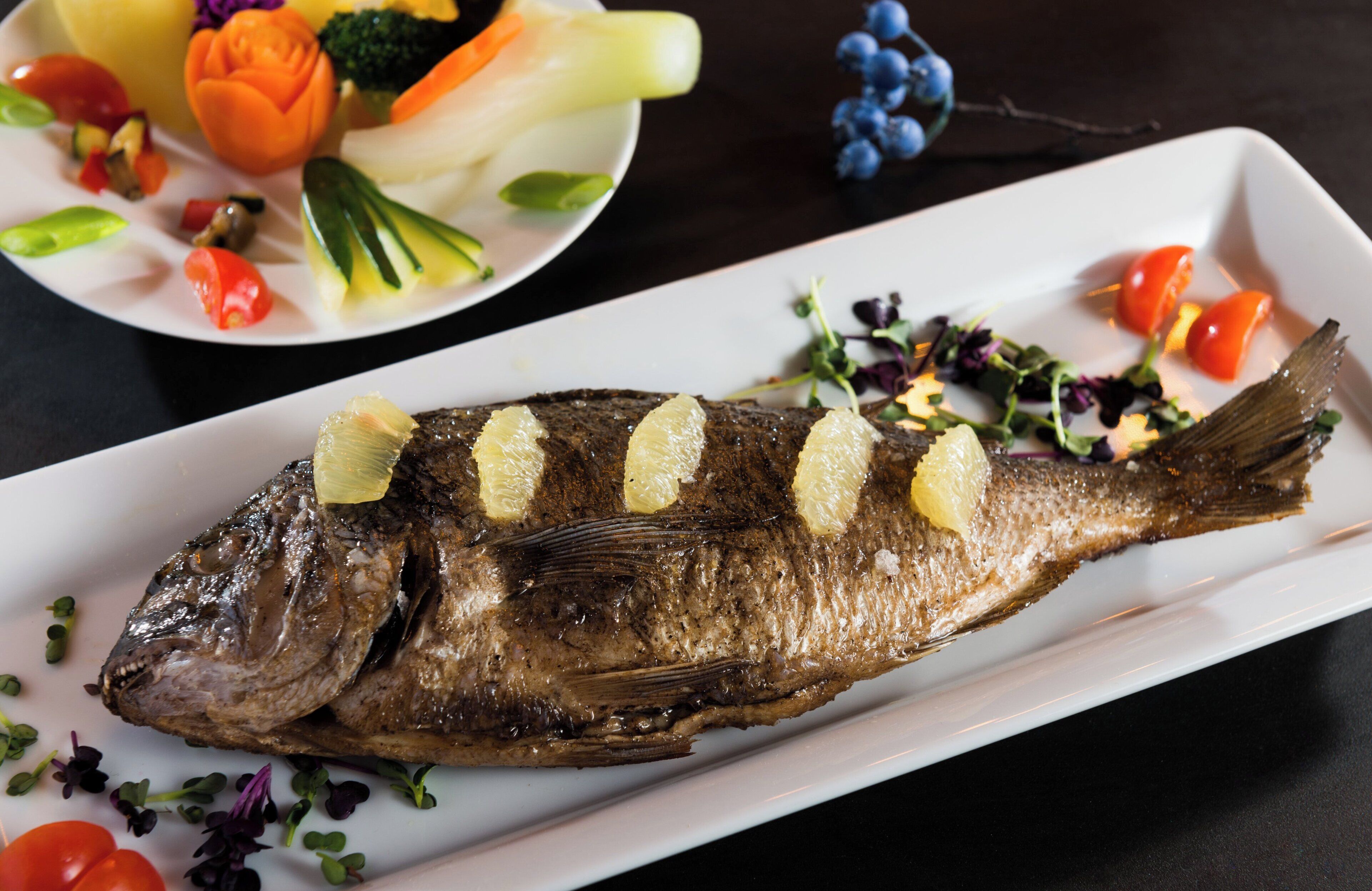Un plato rectangular blanco con un pescado a la parrilla, adornado con rodajas de limón.