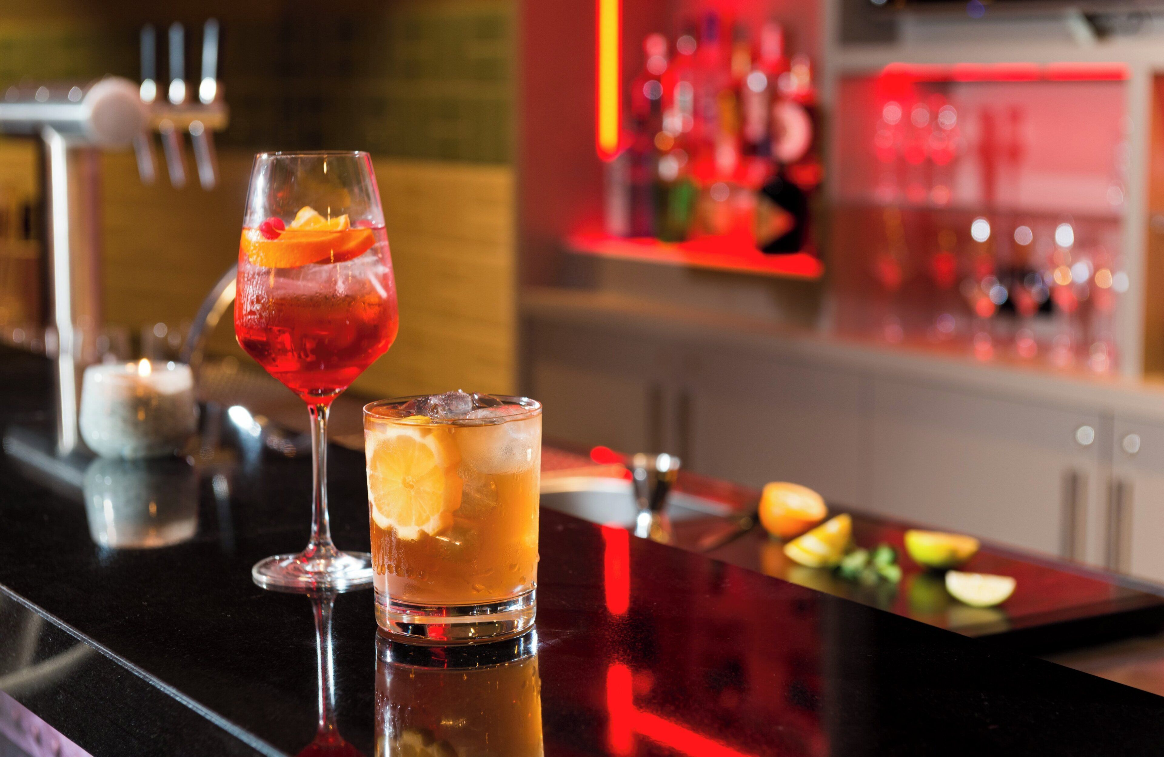 Eine Bar mit zwei Gläsern Getränke, eines mit rotem Inhalt und das andere mit orangefarbenem, sowie einer Auswahl an Flaschen im Hintergrund.