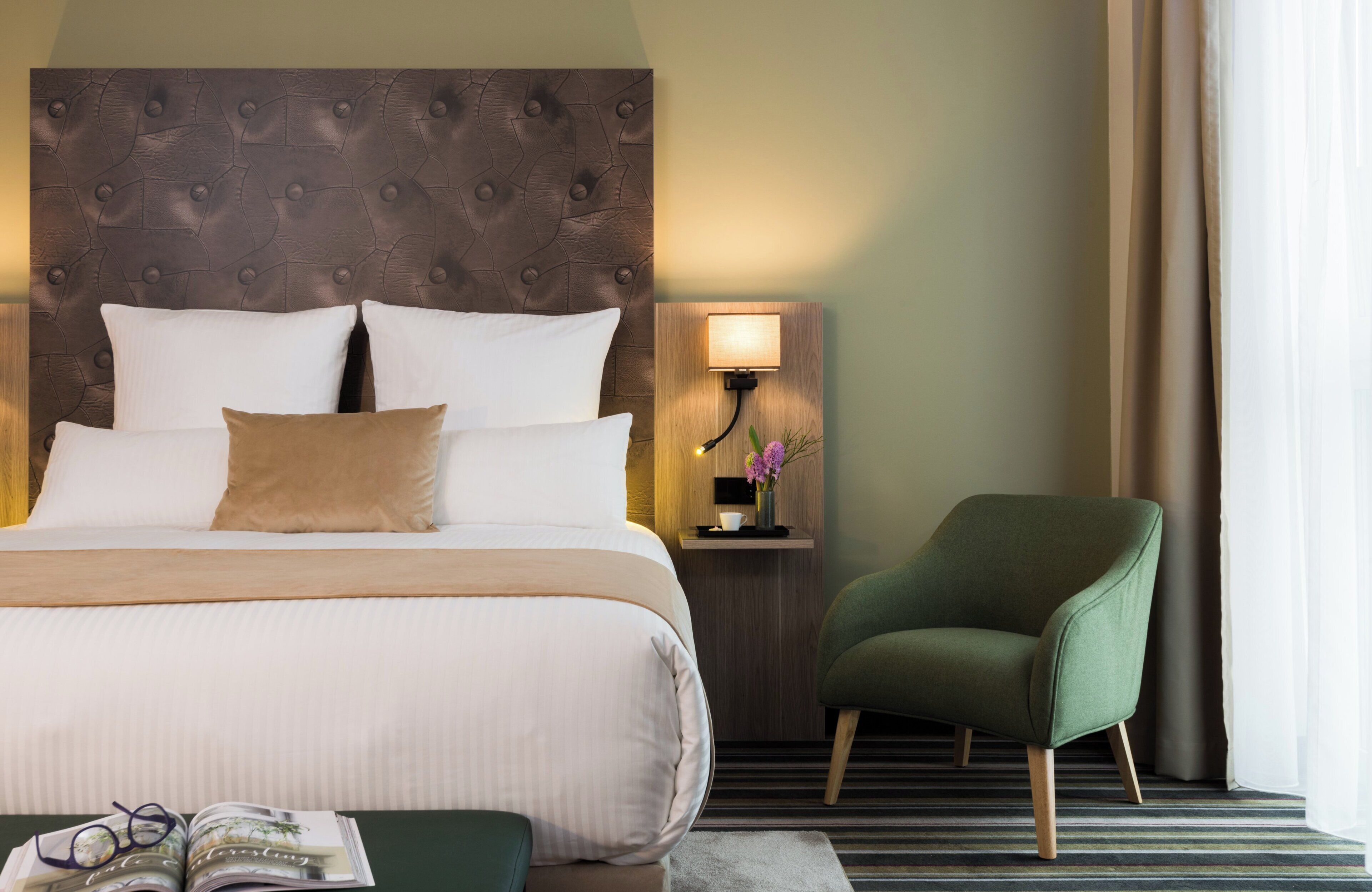 Une chambre d’hôtel avec un grand lit, une table de chevet, un fauteuil vert et une petite table de nuit avec une lampe et des fleurs.